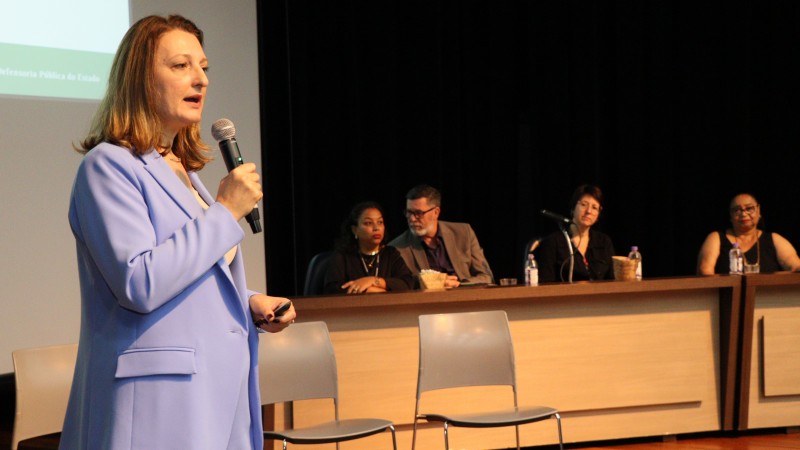 defensora pública, em primeiro plano, falando e, ao fundo, a mesa de abertura do evento, com os convidados assistindo