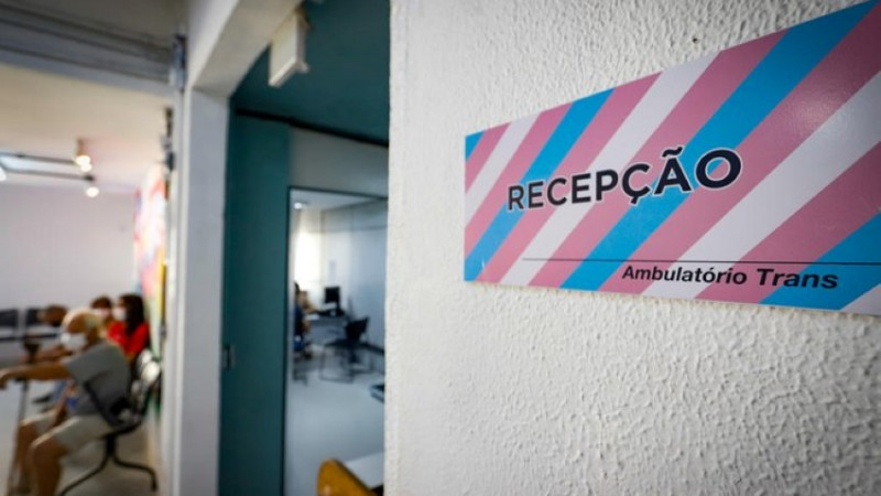 imagem da recepção do ambulatório trans, com a placa escrito "recepção" em primeiro plano e à direita da foto. Ao fundo, algumas pessoas em grupo, e desfocadas