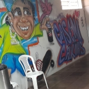 Na foto, vê-se uma parede branca com um grafite de uma menino andando de skate e com uma lata de tinta na mão como se estivesse grafitando as palavras case caxias