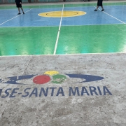 Na foto, vê-se parte da quadra de esportes. Jovens aparecem ao fundo jogando futebol