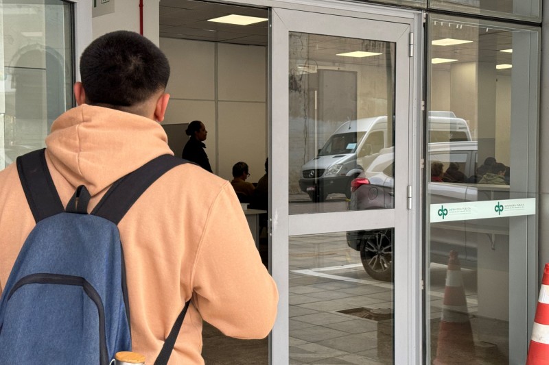 Em primeiro plano, um homem, de costas, está virado para uma porta aberta de entrada a uma sala.