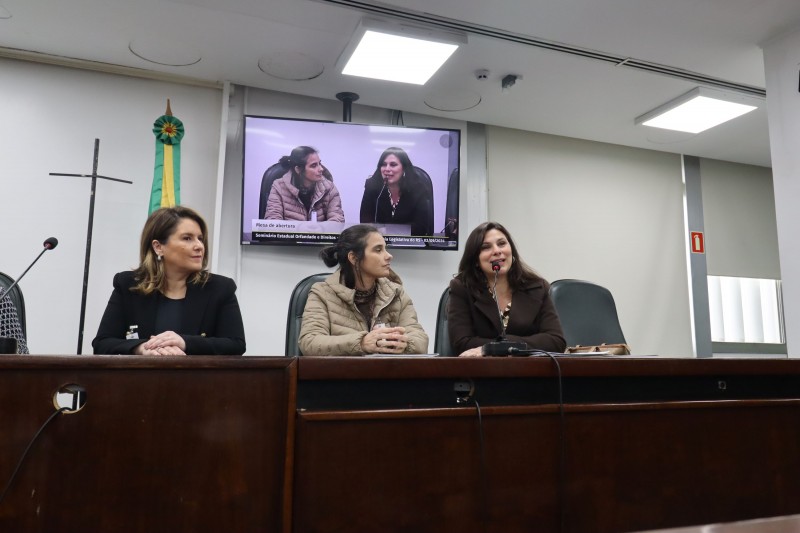 Três mulheres estão sentadas em uma larga bancada. Uma delas, mais à direita, fala ao microfone.