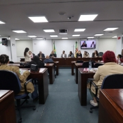 É possível ver fileiras de mesas largas com pessoas sentadas a elas, de costas para a foto. Ao fundo, uma linha de pessoas está sentada a uma mesa maior, virada de frente para a imagem. 