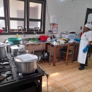 Foto de cozinha com panelas em um fogão, alimentos e bacias em uma pia, mesas e uma cozinheira.