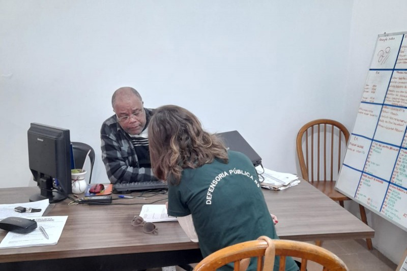 Uma mulher, de costas para a imagem, conversa com um homem que está de frente para a foto. Ambos estão sentados a uma mesa com um computador e papéis.