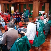 Uma grande sala é vista, com cadeiras vermelhas e piso e pilares em madeira. Há diversas pessoas sentadas, aguardando atendimento, enquanto outras são atendidas por servidores e defensores públicos