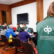 Na foto, em primeiro plano, há uma pessoa de costas, com colete da Defensoria Pública. Ao fundo, desfocado, há pessoas sendo atendidas por servidores e defensores públicos