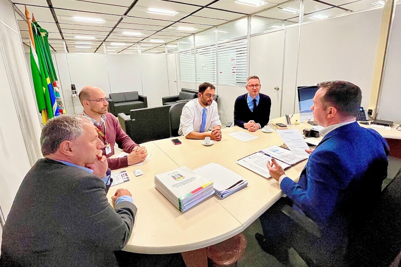 Cinco homens estão sentados a uma mesa dentro de uma sala. Um deles, em primeiro plano, aparece de lado e falando enquanto os outros quatro escutam.