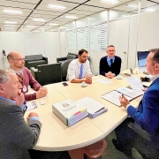 Cinco homens estão sentados a uma mesa dentro de uma sala. Um deles, em primeiro plano, aparece de lado e falando enquanto os outros quatro escutam.