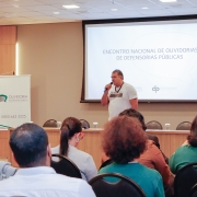 Em segundo plano, um homem de pé com um microfone fala para uma plateia, que aparece em primeiro plano na imagem. Atrás do homem que fala, é possível ver uma mesa de palestra e uma projeção na parede escrito "Encontro Nacional de Ouvidorias de Defensorias Públicas". Também há uma câmera em um tripé, gravando direcionada ao homem de pé.