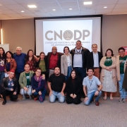 21 pessoas, algumas agachadas no chão e outras de pé, estão juntas posando para uma foto em uma sala. Atrás deles, projetado na parede, há uma lâmina escrito "Conselho Nacional de Ouvidorias das Defensorias Públicas"