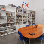 Foto da biblioteca da PMEI. Uma sala branca, com estantes de livros e uma grande mesa no centro, com cadeiras