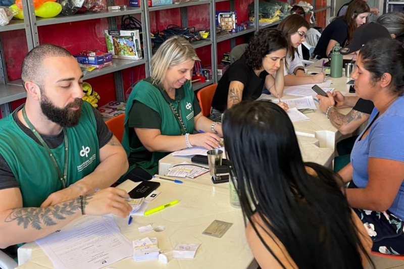 Na foto, há uma sequência de mesas alinhadas, de um lado das mesas sendo possível enxergar duas pessoas com coletes da Defensoria Pública atendendo duas mulheres. Ao fundo, outras pessoas não identificadas com a Defensoria Pública também realizam atendimentos.