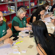 Na foto, há uma sequência de mesas alinhadas, de um lado das mesas sendo possível enxergar duas pessoas com coletes da Defensoria Pública atendendo duas mulheres. Ao fundo, outras pessoas não identificadas com a Defensoria Pública também realizam atendimentos.