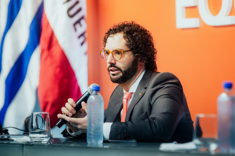 Foto de homem de terno sentado a uma bancada de palestra e falando ao microfone.