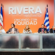 Na foto, é possível ver 6 pessoas sentadas a uma bancada de palestra. Atrás delas, há bandeiras não-hasteadas e um escrito na parede que diz "Rivera sem fronteiras: crescendo com equidade", em espanhol.