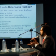 Em primeiro plano da foto e desfocado, é possível ver um homem de terno sentado a uma bancada e falando ao microfone. Em segundo plano e focado na foto, ao fundo, é possível ver uma apresentação de slides projetada na parede com o título "O que é a Defensoria Pública?", em espanhol.