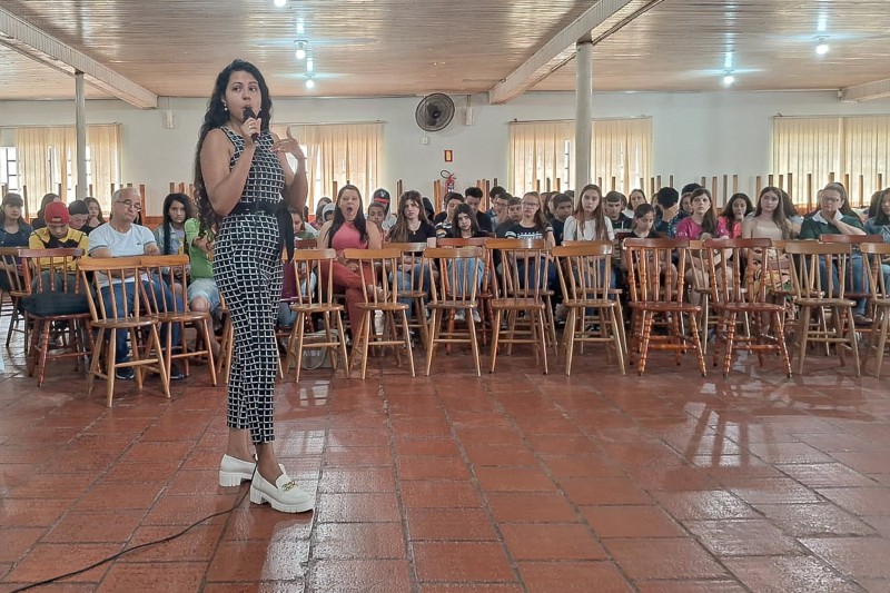 Em primeiro plano, uma mulher está de pé falando a microfone em uma sala, enquanto dezenas de pessoas, em segundo plano e sentadas em cadeiras, escutam.