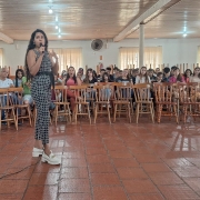 Em primeiro plano, uma mulher está de pé falando a microfone em uma sala, enquanto dezenas de pessoas, em segundo plano e sentadas em cadeiras, escutam.