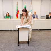 Mulher assinando papel em pequena mesa. Ao fundo, uma bancada com 4 pessoas.