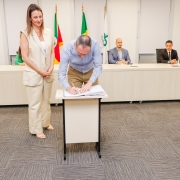 Homem assinando papel em pequena mesa ao lado de mulher que está de pé. Ao fundo, uma bancada com 4 pessoas.