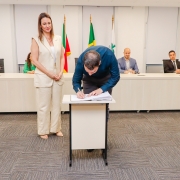 Homem assinando papel em pequena mesa ao lado de mulher que está de pé. Ao fundo, uma bancada com 4 pessoas.