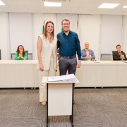 Homem ao lado de mulher posando pra foto, ambos de pé e atrás de pequena mesa com papel. Ao fundo, uma bancada com 4 pessoas aplaudindo.