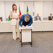 Homem assinando papel em pequena mesa ao lado de mulher que está de pé. Ao fundo, uma bancada com 4 pessoas.