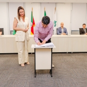 Homem assinando papel em pequena mesa ao lado de mulher que está de pé. Ao fundo, uma bancada com 4 pessoas.