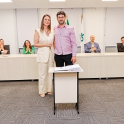 Homem ao lado de mulher posando pra foto, ambos de pé e atrás de pequena mesa com papel. Ao fundo, uma bancada com 4 pessoas aplaudindo.