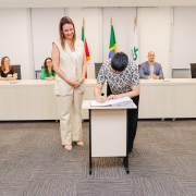 Mulher assinando papel em pequena mesa ao lado de outra mulher que está de pé. Ao fundo, uma bancada com 4 pessoas.