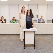 Mulher ao lado de outra mulher posando pra foto, ambos de pé e atrás de pequena mesa com papel. Ao fundo, uma bancada com 4 pessoas aplaudindo.