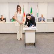 Mulher assinando papel em pequena mesa ao lado de outra mulher que está de pé. Ao fundo, uma bancada com 4 pessoas.