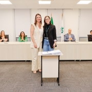 Mulher ao lado de outra mulher posando pra foto, ambos de pé e atrás de pequena mesa com papel. Ao fundo, uma bancada com 4 pessoas aplaudindo.