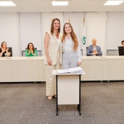 Mulher ao lado de outra mulher posando pra foto, ambos de pé e atrás de pequena mesa com papel. Ao fundo, uma bancada com 4 pessoas aplaudindo.