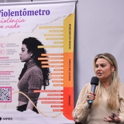 Uma mulher fala ao microfone. Ao lado dela, um cartaz com um infográfico tem o título "Violentômetro: violência se mede" e uma imagem de uma mulher de perfil, além de outros dados e escritos.