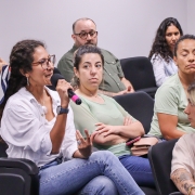 Uma mulher, em meio a uma plateia, fala ao microfone enquanto as pessoas ao redor olham para ela.