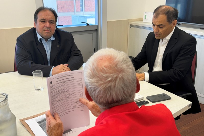 Em primeiro plano, um homem de costas para a imagem segura um papel sobre uma mesa. Em segundo plano, dois homens de frente para a imagem e de frente para o outro homem estão sentados à uma mesa o escutando