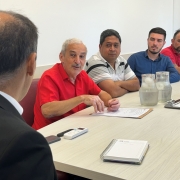 Em primeiro plano, Um homem, de costas para a foto, está sentado a uma mesa. Em segundo plano, outros 4 homens, de frente para a foto, estão sentados na mesma mesa e um deles está falando com o homem que está de costas.