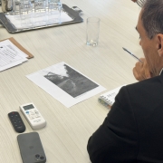 Um homem, de costas para a imagem, segura uma caneta e olha para um papel com uma foto em cima de uma mesa.