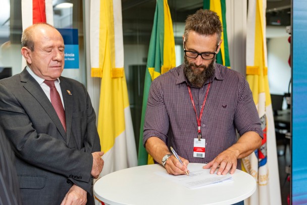 Homem assinando papel em mesa ao lado de outro homem que olha para o papel sendo assinado.