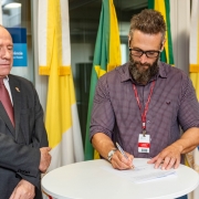 Homem assinando papel em mesa ao lado de outro homem que olha para o papel sendo assinado.