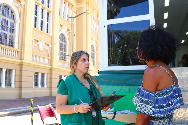 Na rua uma mulher de costas para a câmera fala com outra mulher que, com uma prancheta e uma caneta, escuta o que a primeira mulher fala