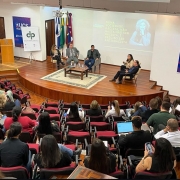 Vista superior de um auditório lotado, com um palco composto por 5 pessoas. Dentre elas, Alessandra Quines, defensora pública. 