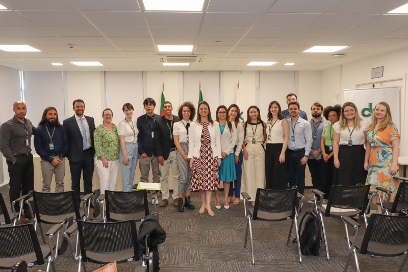 19 pessoas em uma sala, todas de pé, uma do lado da outra, posando para foto.