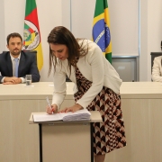 Mulher assinando papel em pequena mesa. Ao fundo, uma mulher e um homem estão sentados a uma mesa e olhando para ela. 