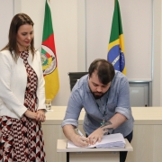 Foto de homem assinando papel em pequena mesa enquanto uma mulher, de pé e ao seu lado, observa a assinatura.