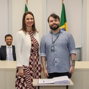 Um homem e uma mulher, de pé e um ao lado do outro, posam para foto atrás de uma mesa com um papel e uma caneta.