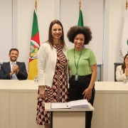 Duas mulheres, de pé e uma ao lado da outra, posam para foto atrás de uma mesa com um papel e uma caneta.