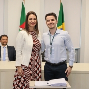 Um homem e uma mulher, de pé e um ao lado do outro, posam para foto atrás de uma mesa com um papel e uma caneta.